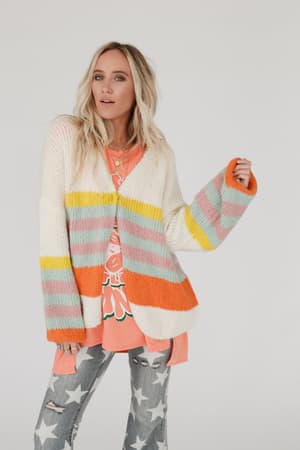 A woman with blonde hair wears the Retro Babe Striped Cardigan - Cream over a bright graphic tee and ripped star jeans. Against a light background, she looks at the camera with a relaxed expression.
