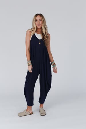 A person with long blonde hair wears The Perfect Harem Jumpsuit in navy over a white top, with tan slip-on shoes, layered necklaces, and multiple bracelets, standing against a plain light background.