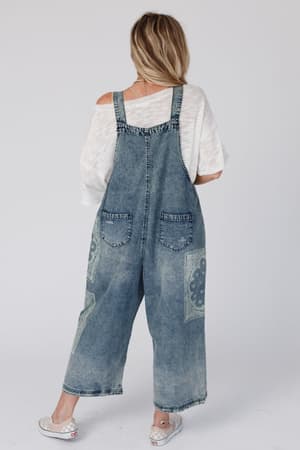 A person with long, wavy blonde hair stands facing away, wearing the High Hopes Slouchy Denim Overalls - Denim Bandana over a white off-shoulder top and white sneakers against a plain light background.