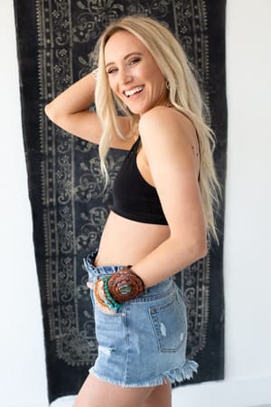 A smiling woman with long blonde hair poses sideways, wearing the Floral Cut Out High Neck Bralette in black, paired with denim shorts and colorful bracelets, standing in front of a dark patterned wall hanging.