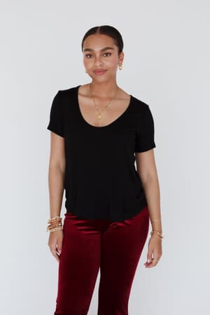 A woman with dark hair in a low bun wears the Scoop Neck Bralette Tee in black, paired with red velvet pants and gold jewelry, standing against a plain white background and smiling slightly.