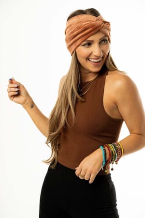 A smiling woman poses against a white background wearing the High Neck Racerback Brami in brown, paired with black pants, a rust-colored headband, and colorful bracelets on her right arm.