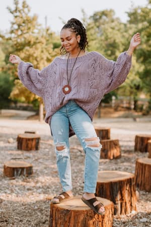 A woman standing outdoors on a tree stump, smiling with arms raised, wears the Golden Hour Bubble Sleeve Sweater Dress in Lavender, surrounded by greenery and more stumps.