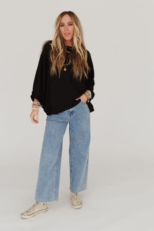 A woman with long blonde hair wears the Fly With Me Batwing Sleeve Knit Sweater in black, paired with light blue flared jeans, white sneakers, and layered jewelry. She stands against a plain white background, looking at the camera.
