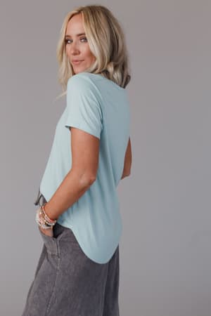 A woman with long blonde hair models the Scoop Neck Bralette Tee in Washed Seafoam, paired with gray pants. She stands sideways, smiling over her shoulder, wearing bracelets against a plain gray background.
