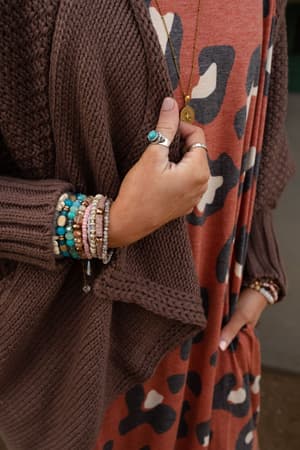 A person wears a brown knit cardigan over a rust leopard-print dress, accessorized with layered beaded bracelets, boho jewelry, and the Kapalua Bay Adjustable Turquoise Ring - Silver plus a long pendant necklace.