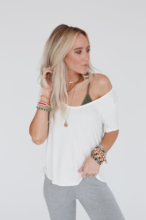 A woman with long blonde hair wears the Scoop Neck Bralette Tee in white over a green bralette and gray leggings, accessorized with colorful bracelets and a necklace, looking to the side against a plain white background.