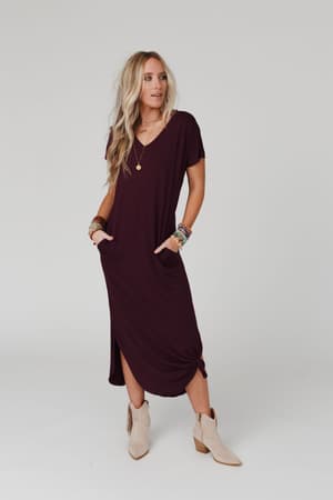 A woman with long blonde hair models the Call It Comfort Tee Dress in plum, a boho relaxed-fit maxi with pockets, paired with tan ankle boots, layered bracelets, and a gold necklace against a plain light background.
