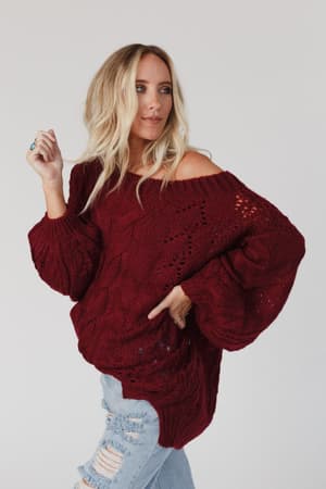 A woman with long blonde hair wears the Golden Hour Bubble Sleeve Sweater Dress in burgundy, styled off one shoulder, and poses against a plain white background.
