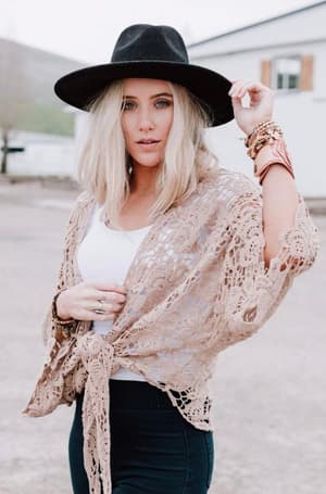 A blonde woman wearing a wide-brim black hat, white top, and the Crochet Petal Kimono Wrap in Mocha poses outdoors with stacked bracelets, holding her hat brim—a true boho closet inspiration.
