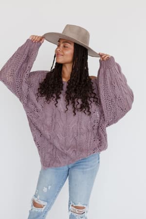 A woman stands against a plain white background, smiling with her eyes closed and holding her hat while wearing the Golden Hour Bubble Sleeve Sweater Dress in lavender and a wide-brimmed gray hat.