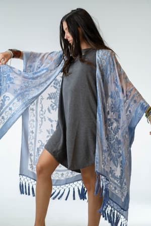 A smiling woman with long dark hair poses in a loose grey dress, showcasing the Crushin' On You Velvet Kimono in light blue with tasseled edges against a plain light background.