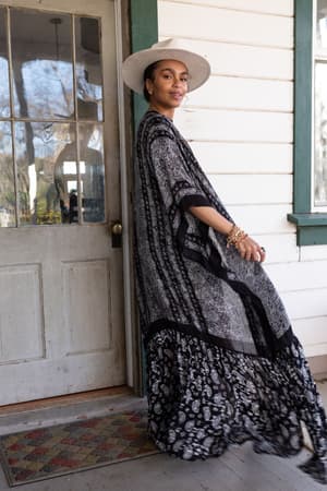 A woman in a wide-brimmed hat and the Paisley Tapestry Free Flow Duster Kimono - Black stands on a porch, smiling as she turns toward the camera. Behind her are a glass-paneled door and window.