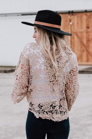 A woman with long blonde hair stands with her back to the camera, wearing a black hat and the Crochet Petal Kimono Wrap - Mocha, paired with dark jeans. She poses outdoors near a wooden door, capturing that effortless boho closet vibe.