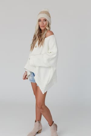 A woman in the Dana Oversized Diamond Texture Sweater - Ivory, denim shorts, a cream knit beanie, and beige ankle boots stands against a plain light background, looking at the camera with a relaxed expression.