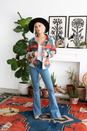 A woman in a black hat, white top, blue flared jeans, and the Anika Mixed Pattern Jacket - Teal stands indoors on a colorful rug, surrounded by potted plants, framed artwork, and a decorative skull on the mantel.