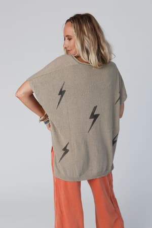 A blonde woman faces away, wearing the Struck By You Dolman Sleeve Sweater Top in mocha with black lightning bolt patterns and loose orange pants. The background is plain and light-colored.