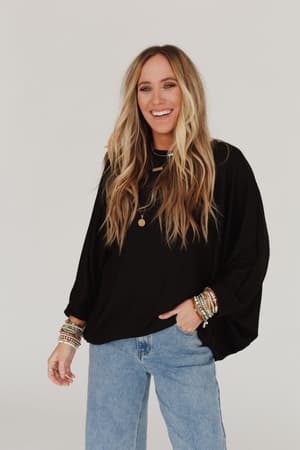 A woman with long, wavy blonde hair wears the Fly With Me Batwing Sleeve Knit Sweater in black, paired with blue flared jeans, layered necklaces, and bracelets. She stands against a white background, smiling with one hand in her pocket.