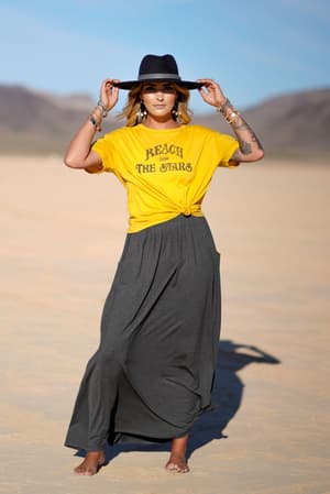 A woman stands barefoot on a sandy plain, wearing a yellow Reach for the Stars t-shirt, The Perfect Pocketed Maxi Skirt in Charcoal, jewelry, and a black hat, with mountains and blue sky in the background.