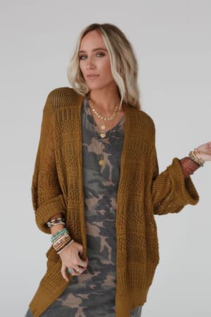 A woman with blonde hair poses confidently against a plain light background wearing a camouflage dress, layered necklaces, multiple bracelets, and the Brenden Knit Netted Cardigan in brown.