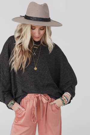 A woman in The Nest Fly With Me Batwing Sleeve Knit Sweater in charcoal, pink drawstring pants, and a wide-brimmed hat looks down. She accessorizes with layered necklaces, colorful bracelets, and her long blonde hair is loosely waved.