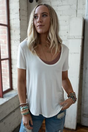 A woman with long blonde hair stands indoors by a window with a white brick wall, wearing the Scoop Neck Bralette Tee in white over a mauve camisole, paired with distressed blue jeans and beaded bracelets.