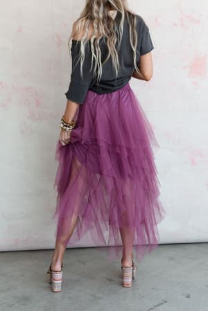 A person with long, wavy hair poses by a light wall in a dark top and the Boho Ballerina Skirt - Mauve. Lifting the layered tulle skirt, they show high-heeled sandals, creating a casual and playful look.