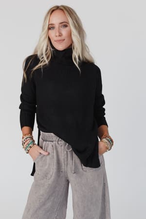 A woman with long blonde hair wears the Danny Sweater - Black, paired with gray wide-leg pants and colorful beaded bracelets. With her hands in her pockets, she looks confidently at the camera against a plain background.