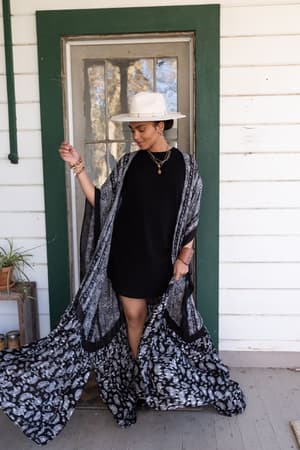 A woman in a white hat, black dress, and the Paisley Tapestry Free Flow Duster Kimono - Black stands on a porch before a green-framed door, looking down with one hand raised. A small plant rests on a nearby shelf.