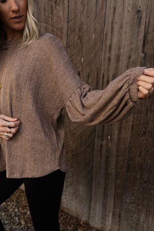 A person wears The Nest Sweet Heart Balloon Sleeve Knit Sweater in Mocha with black pants, standing by a wooden wall and holding out one balloon sleeve to highlight its gathered, puffy design. Their face is partially out of frame.