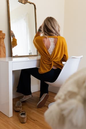 A blonde woman sits at a white vanity in The Nest Flattering Open Back Thermal Knit Top - Mustard, revealing lace details. Facing the mirror with one hand in her hair, she pairs the relaxed boho look with casual shoes.