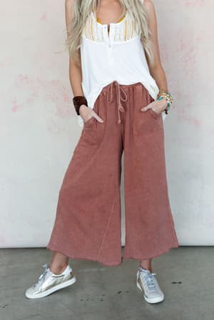 A woman stands against a light wall, wearing a sleeveless white top, Relaxing Robin Wide Leg Pant in Red Bean, metallic sneakers, and colorful beaded bracelets, with her hands in her pockets.