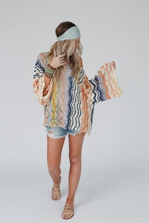 A woman wears the Rainbow Shores Knit Sweater - Multi with its colorful boho style, slouchy fit, and flare sleeves. She pairs it with denim shorts, sandals, a blue headband, and bracelets while standing against a white background.