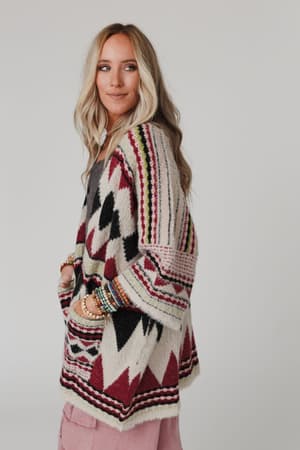A woman with long blonde hair models the Keep It Up Oversized Cardigan - Berry in red, black, cream, and green tones. She stands sideways with hands in pockets, smiling gently. Pink pants and layered bracelets finish her look against a gray background.