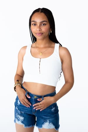 A woman with long braids wears the Hook It Up Ribbed Brami - Ivory and distressed denim shorts, standing against a plain white background and smiling slightly at the camera.