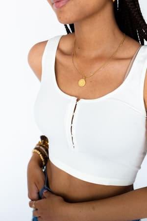 A woman stands with one hand on her hip, wearing the Hook It Up Ribbed Brami in ivory, gold layered necklaces, and bracelets. Her braided hair and part of her face are visible against a white background.