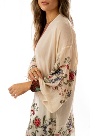 A woman models the Fun In Florals Kimono - Cream, featuring light, flowy fabric with colorful floral embroidery on sheer sleeves and hem. She stands against a white background with loose hair, accessorized with rings and bracelets.