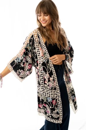 A woman smiles while wearing the Let's Get Tropical Kimono - Black, a long, loose floral print piece with colorful embroidery and white lattice trim, over a black top and dark jeans against a plain white background.