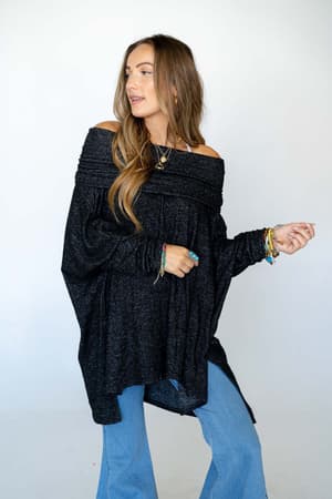 A woman with long brown hair poses against a white background in The Nest So Versatile Flowy Knit Sweater - Charcoal, styled with light blue flared jeans, layered necklaces, rings, and colorful bracelets.