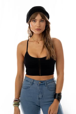A woman with long brown hair wears the Sweet + Seamless Bralette in Black, a black knit beanie, blue high-waisted jeans, layered necklaces, bracelets, and a black wristband as she stands against a plain white background looking at the camera.
