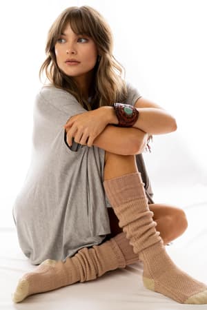 A woman with long, wavy hair sits on the floor in a relaxed pose, wearing Cozy Cabin Lounge Socks - Pink and a loose gray t-shirt. She gazes thoughtfully to the side against a plain white background.