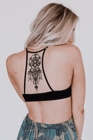 A woman with long blonde hair models the Tattoo Bralette - Black, revealing an intricate black tattoo design on her upper back. She faces away, looking over her shoulder, pairing the bralette with patterned pants for a boho look.