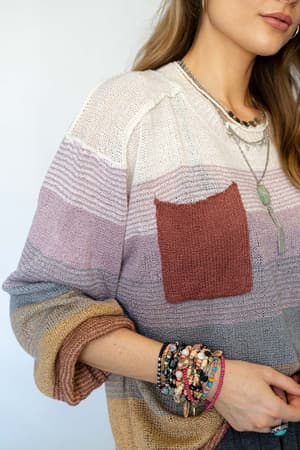 A woman wears the Going Places Gradient Stripe Sweater - Mauve, featuring a red pocket, along with layered necklaces and colorful beaded bracelets. Only her torso and part of her face are visible.