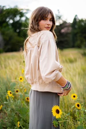 A woman with wavy brown hair stands in a field of yellow wildflowers, wearing the Lorelei Crochet Jacket in beige and a gray skirt, looking over her shoulder at the camera. She accessorizes with colorful bracelets on her wrist.