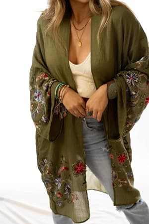 A woman wears the Fun In Florals Kimono - Olive over a cream top with light blue ripped jeans, layered necklaces, and multiple bracelets, standing against a plain white background.