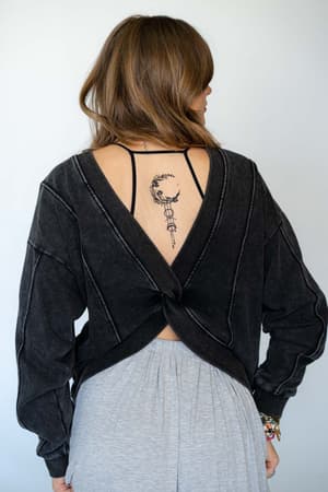 A woman with light brown hair faces away, wearing the Crescent Moon Tattoo Bralette - Black, which has an open back showing her crescent moon and arrow tattoo. She also wears light gray pants and beaded bracelets.