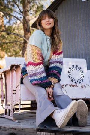 A woman sits on a rustic white bench outdoors, smiling slightly. She wears the Wrapped up in Color Knit Cardigan - Neutral over a light blue shirt, with gray flared pants and white sneakers. Trees and a corrugated metal wall are in the background.