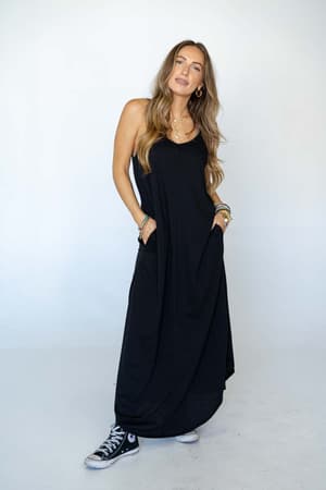 A woman with long wavy hair wears The Nest Perfect Round Hem Bralette Maxi Dress - Black with pockets, pairing it with black Converse sneakers. She stands against a plain light background, hands in her pockets and a calm expression on her face.