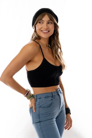 A woman wears the Sweet + Seamless Bralette - Black, high-waisted blue jeans, a black beanie, colorful bracelets, and a necklace as she smiles and looks to the side against a white background.