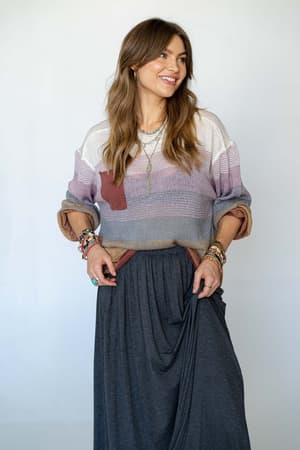 A woman with long brown hair smiles in the Going Places Gradient Stripe Sweater - Mauve and a dark gray maxi skirt, accessorized with layered necklaces and colorful bracelets, standing against a plain light background.